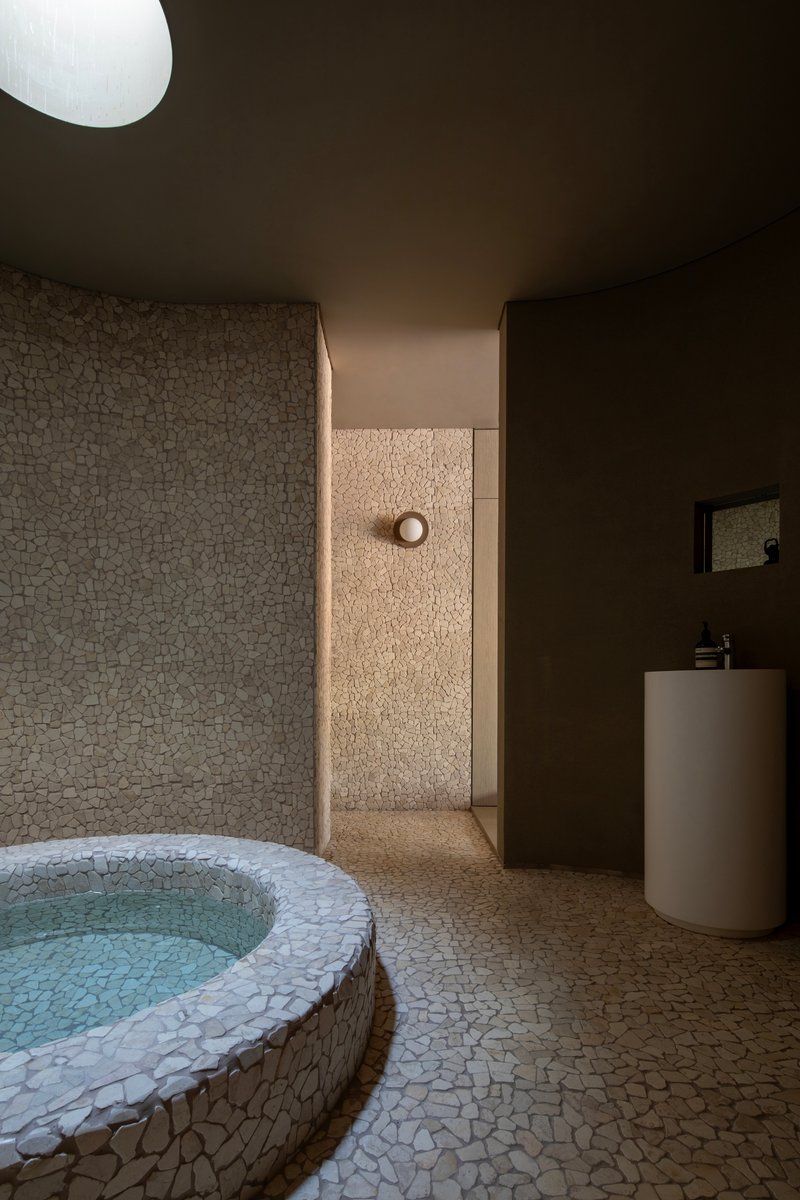 Bathing room with circular stone-clad soaking tub, pebble mosaic walls and cylindrical white basin under skylight