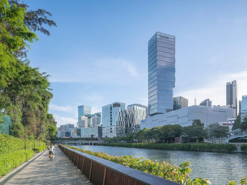 Waterfront pathway with cyclist beneath trees looking toward glazed tower and skyline beyond