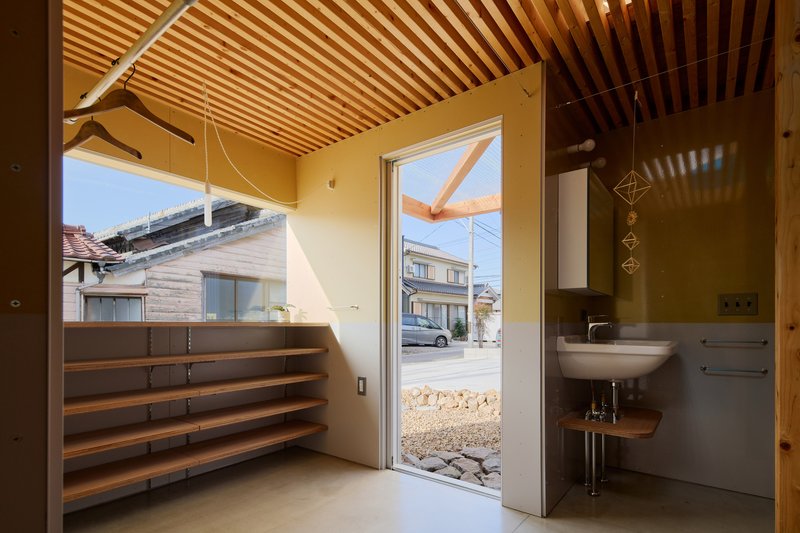 Entry corner with timber shelving and wall-mounted sink opening to a gravel courtyard and neighboring houses