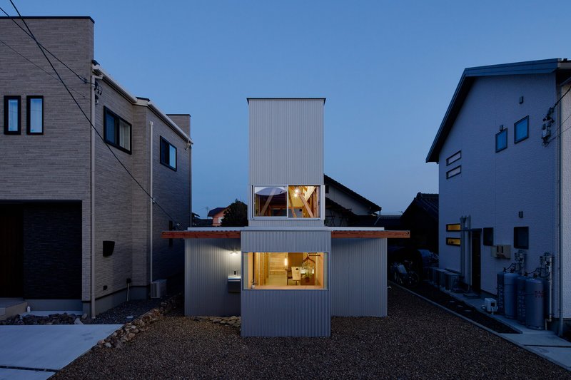 Corrugated metal clad volume with illuminated windows at dusk nestled between neighboring houses in gravel yard