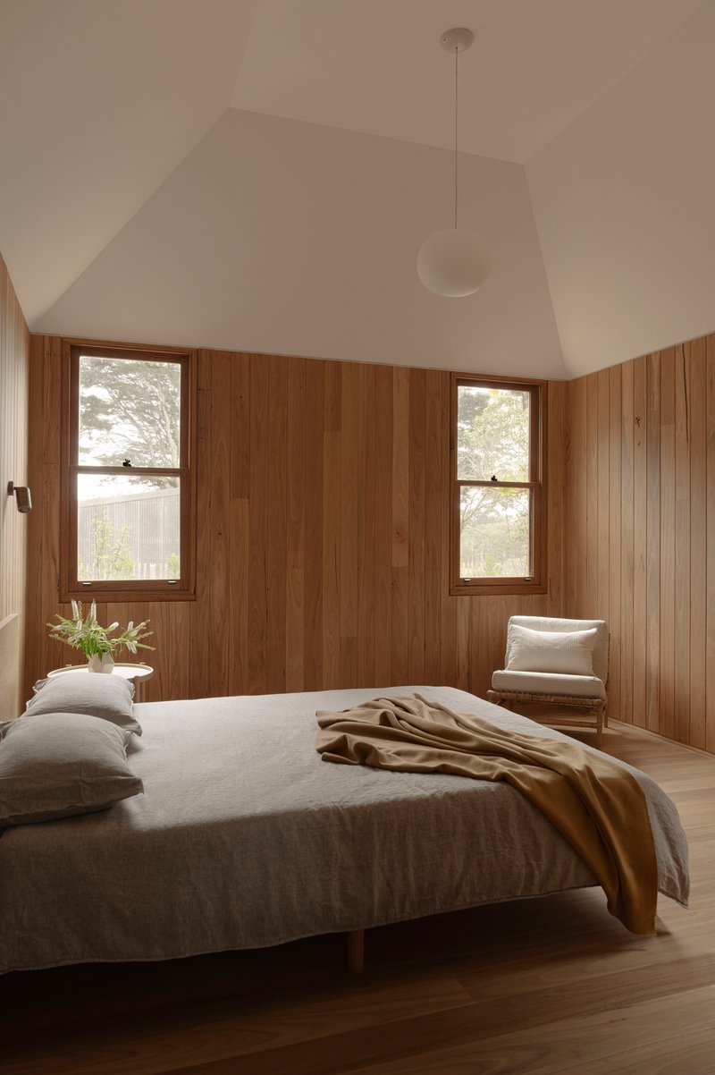 Bedroom with vertical timber paneling, vaulted white ceiling, and two framed windows flanking the bed