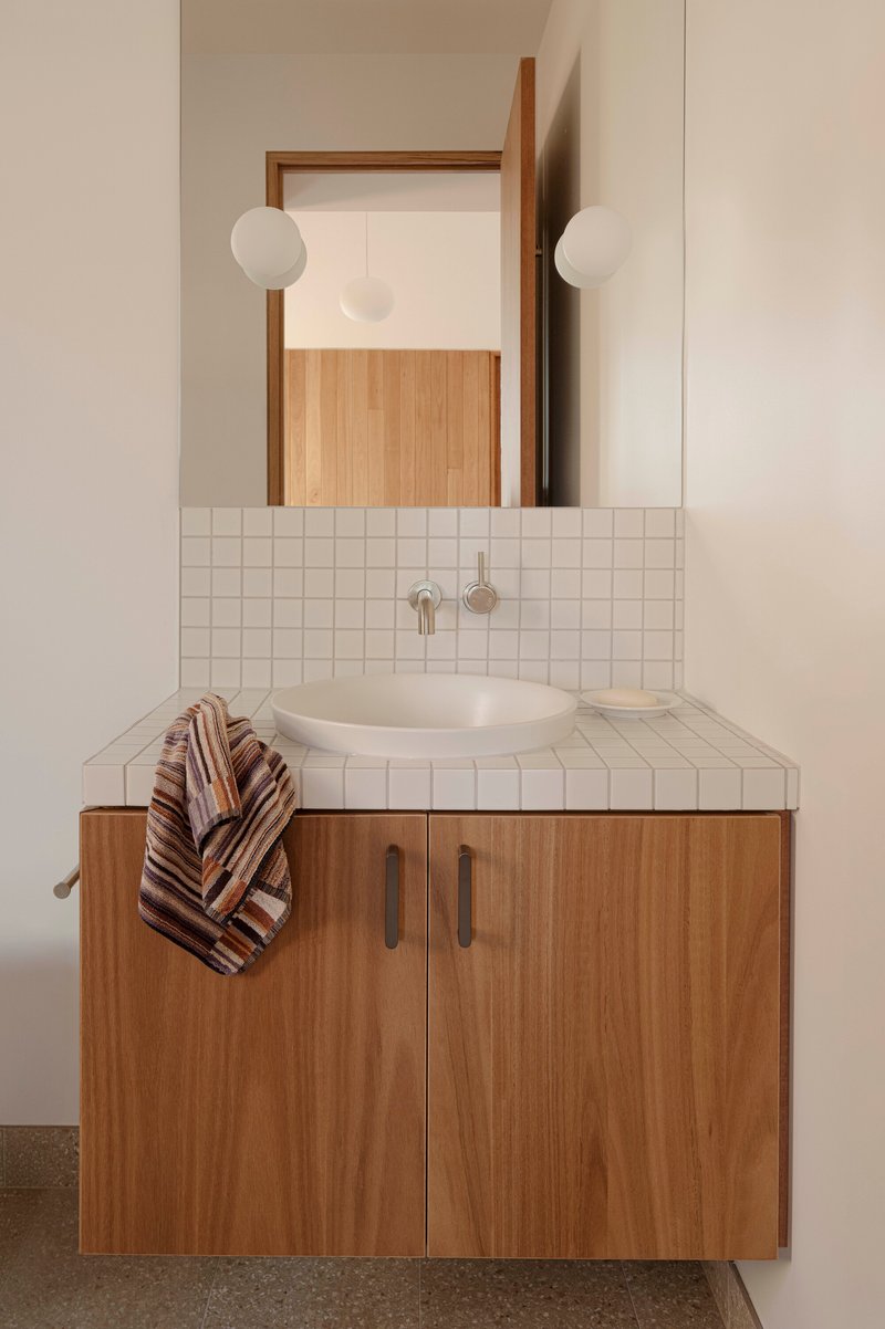 Bathroom vanity with white square tile counter, timber cabinetry and twin globe wall sconces flanking a mirror