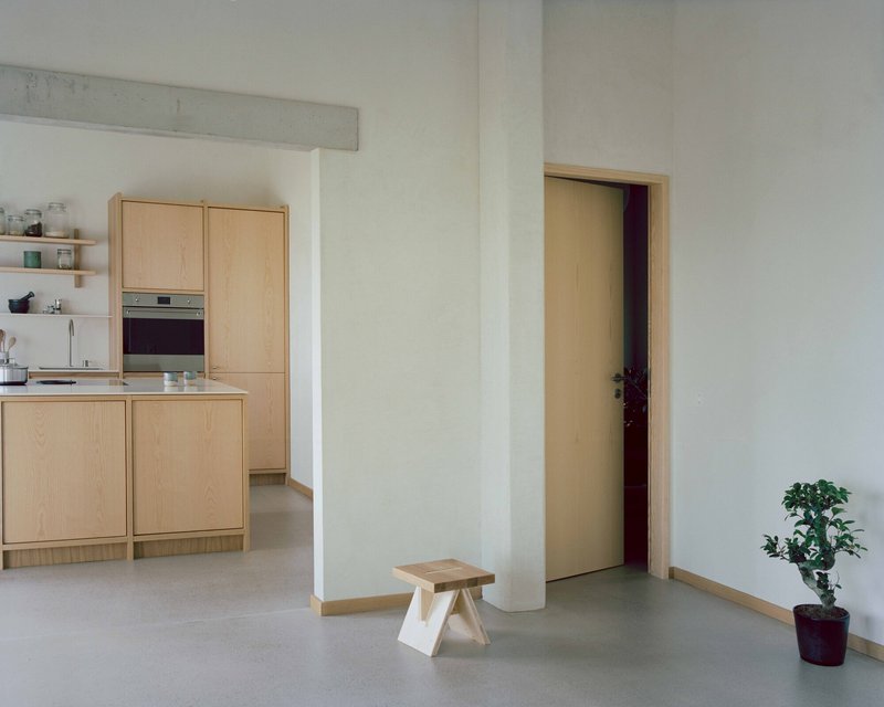 White-walled corridor with timber-framed doorway and small wooden stool beside a potted plant
