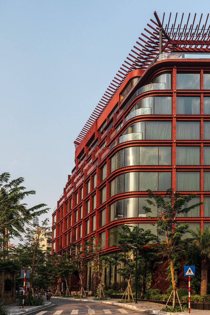 Curved red metal facade with horizontal louvers and brise-soleil crown at street level with young trees