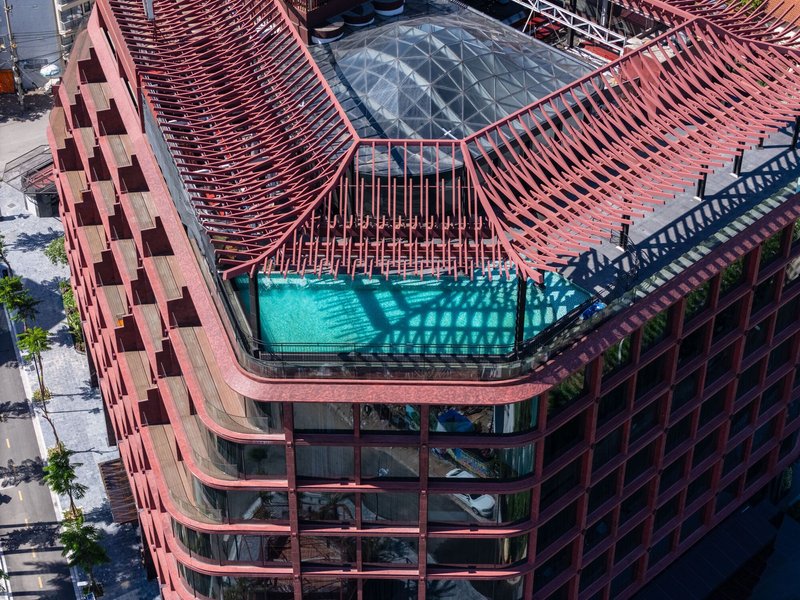 Aerial view of rooftop pool beneath red metal brise-soleil frame and glazed skylight opening