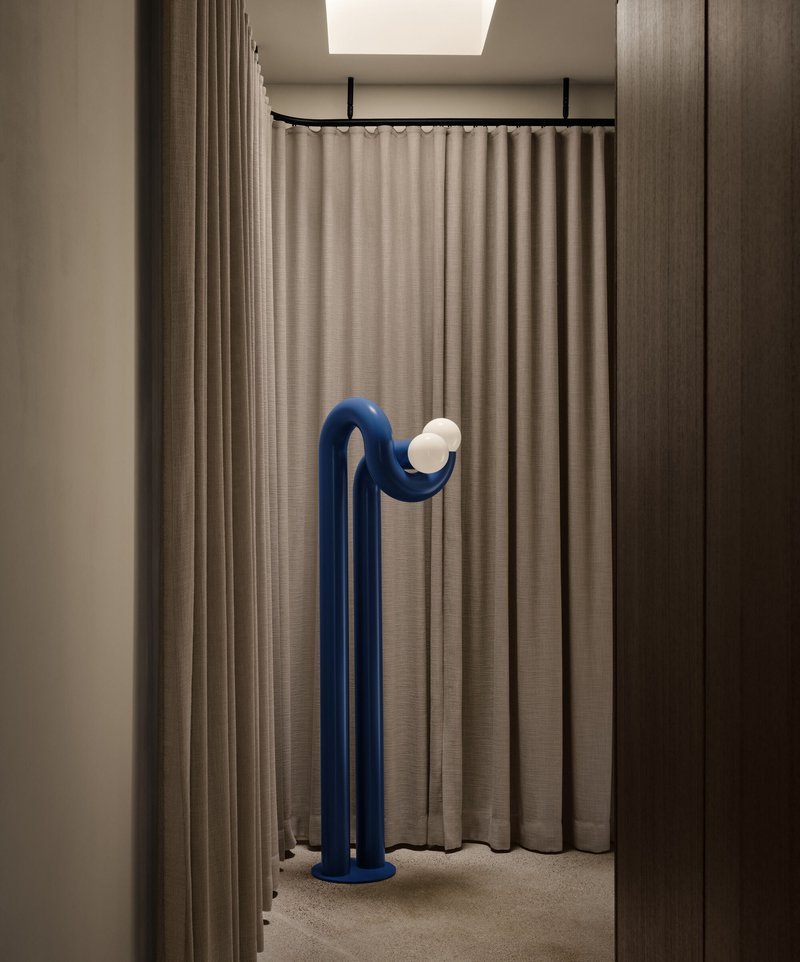Blue tubular floor lamp with spherical bulb standing against beige curtain beneath square skylight