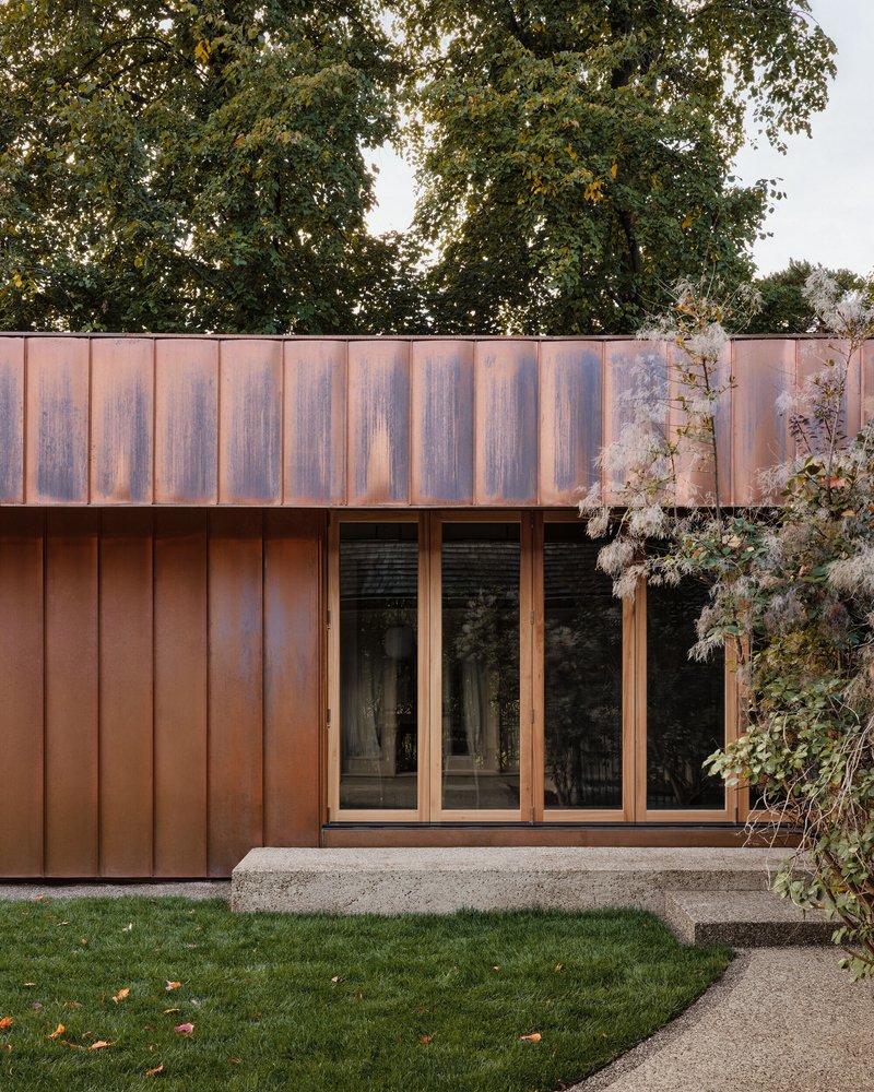 Copper-clad facade with vertical panels and timber-framed glazing adjacent to planted rose bushes