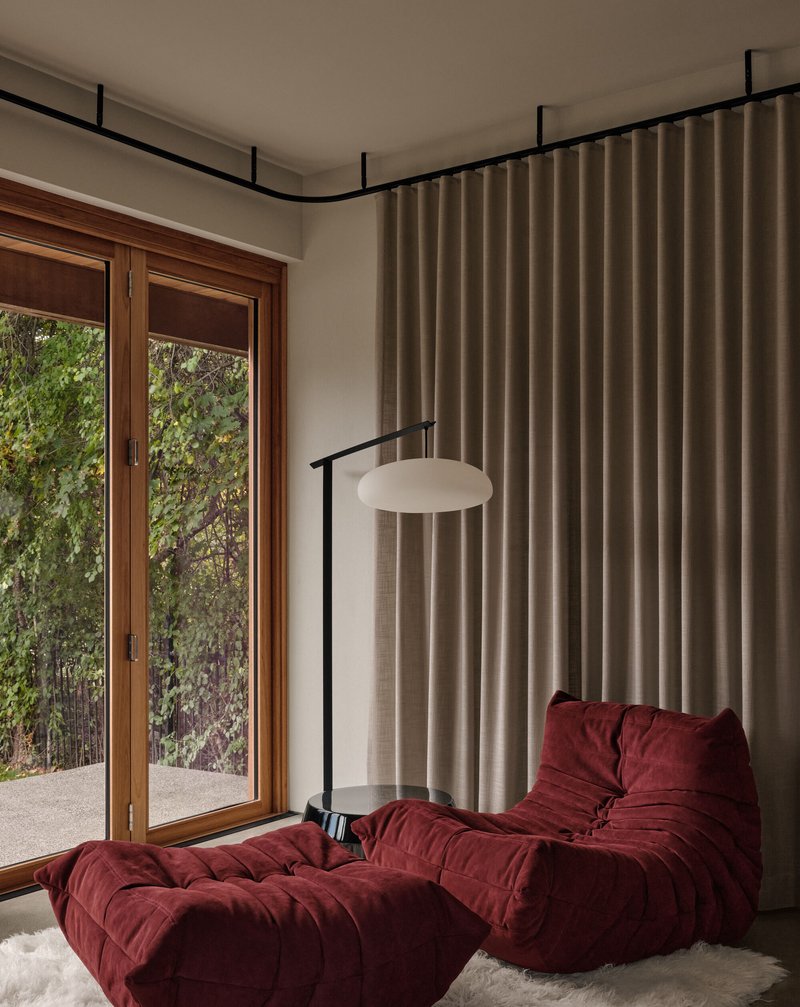 Reading nook with timber-framed glazing, beige curtain track and burgundy upholstered seating overlooking greenery