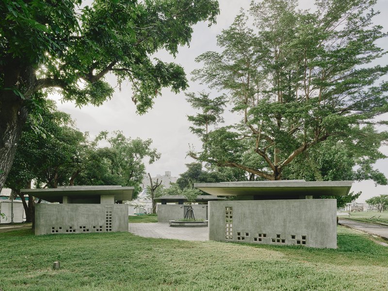 Pair of concrete pavilions with flat roofs and perforated walls beneath mature trees in a park