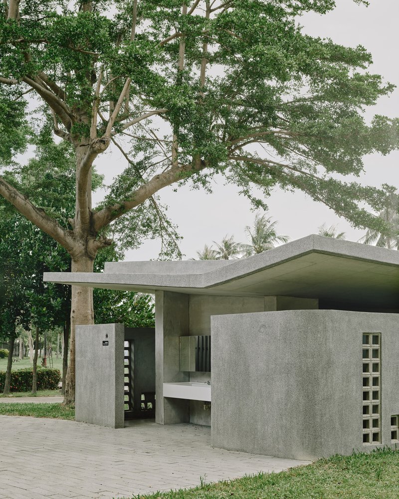 Corner view of concrete pavilion with cantilevered roof plane sheltering a bench beneath tree canopy