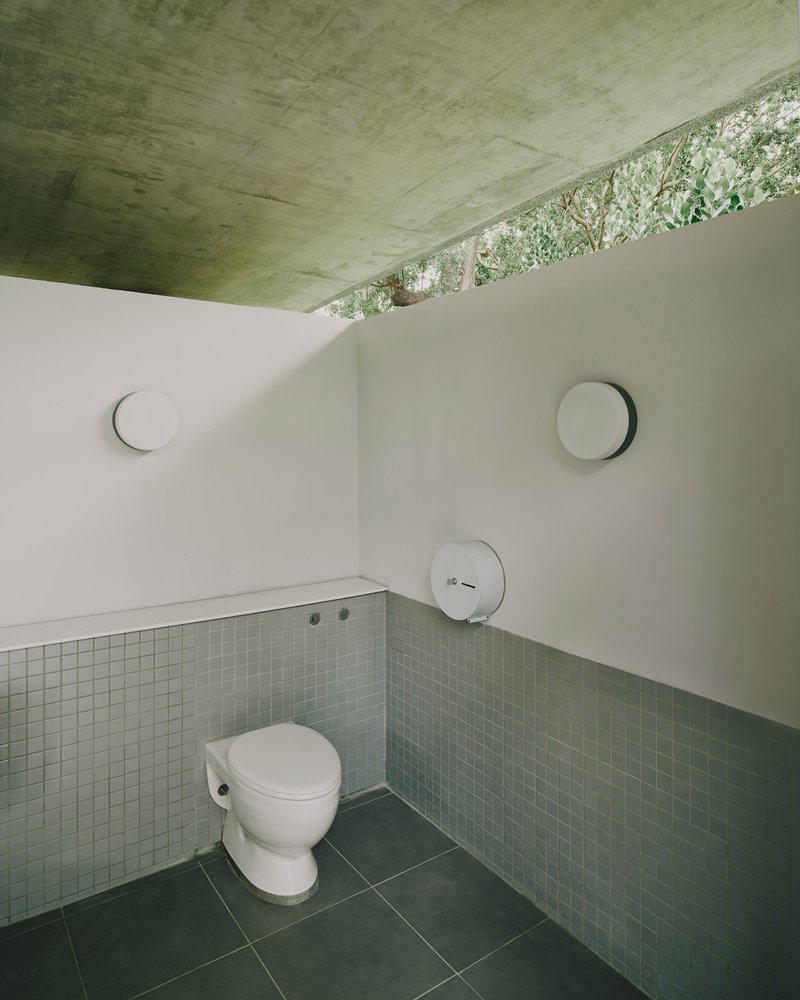 Public restroom interior with green mosaic tile wainscot and concrete ceiling opening to foliage above