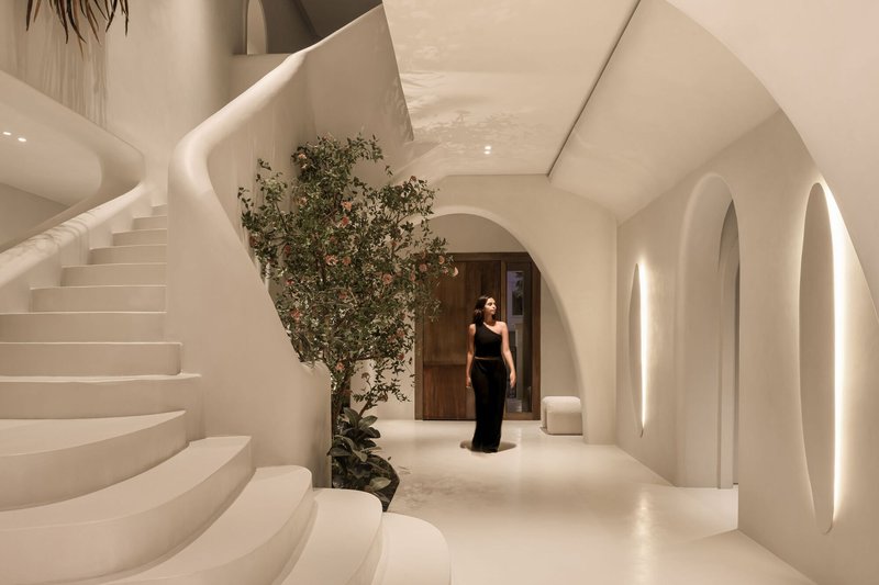 White curved entry hall with spiral staircase and arched doorways, a flowering tree and a visitor in evening light