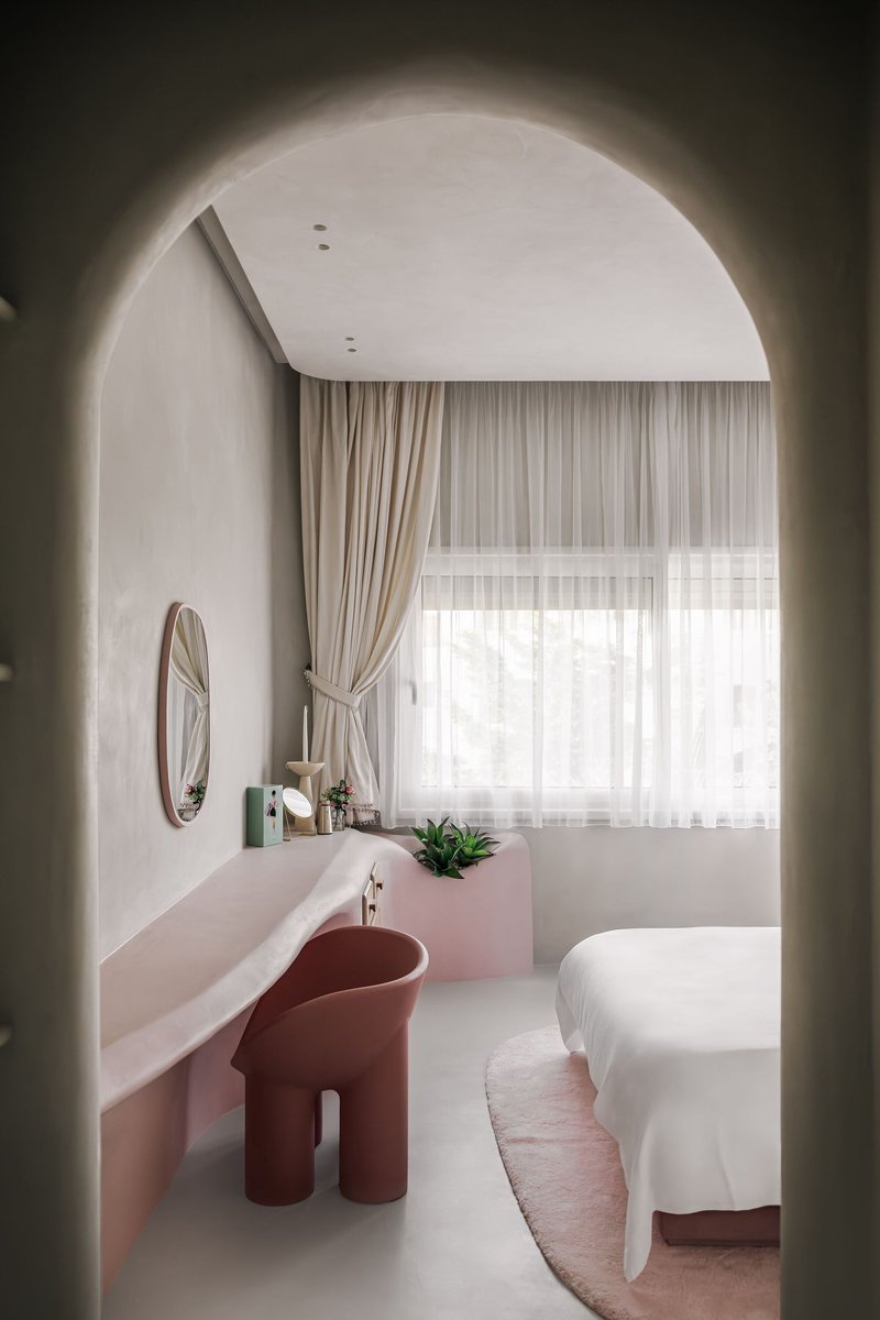 Bedroom viewed through rounded archway with pink ledge desk and terracotta chair facing draped windows