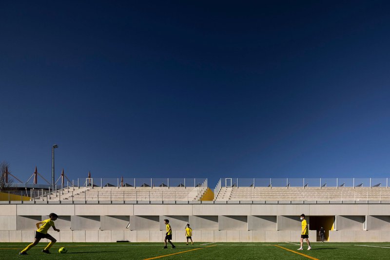 Concrete grandstand with rhythmic column openings and players in yellow passing by on the pitch
