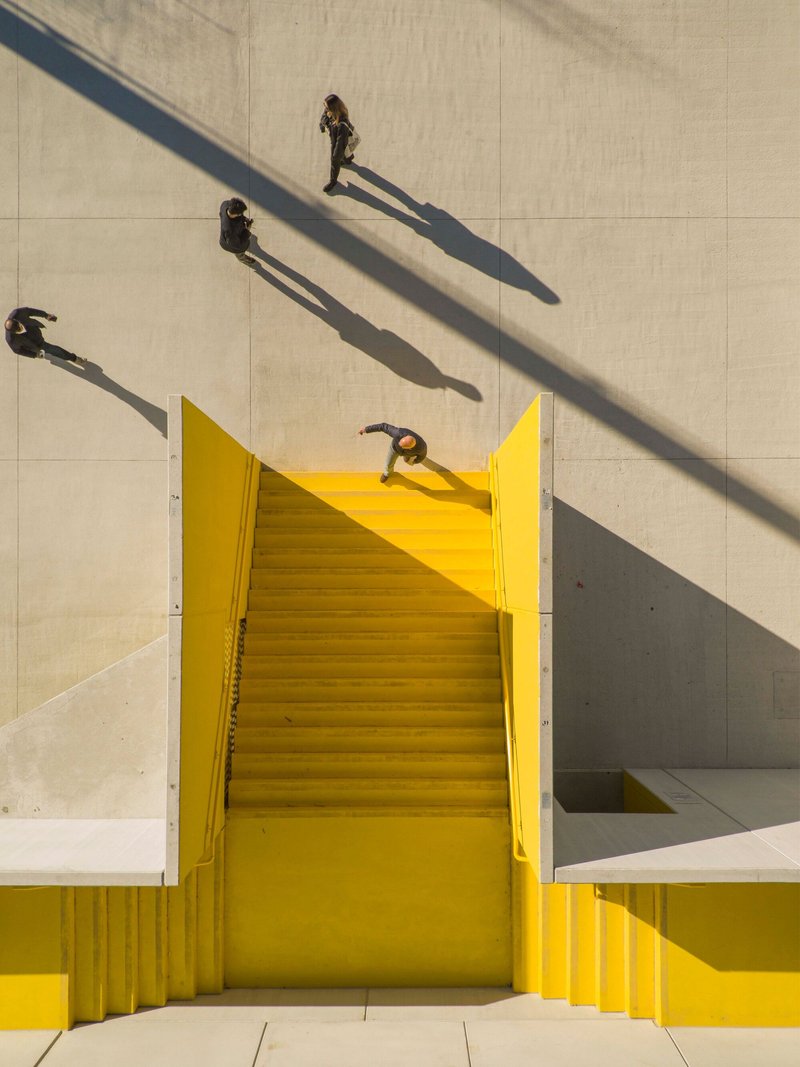 Aerial view of yellow staircase bisecting concrete plaza with pedestrians casting long shadows
