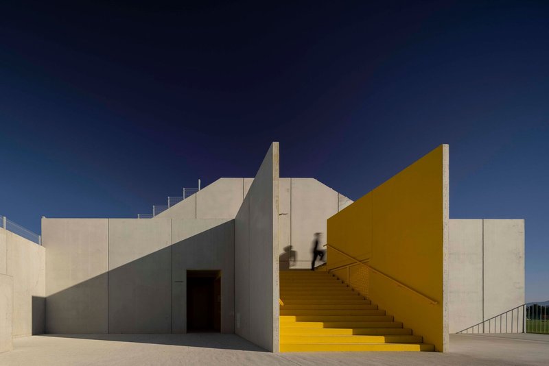 Two figures crossing an elevated walkway between white and yellow building volumes at dusk
