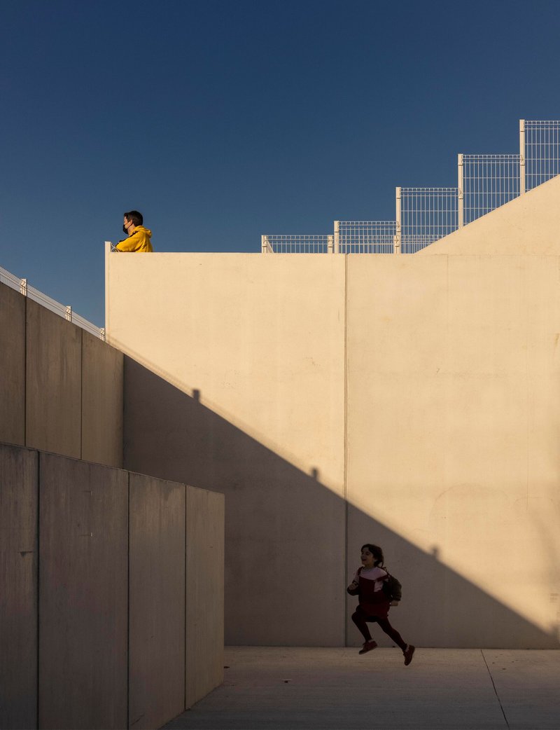 Concrete walls and courtyard with child running across sunlit pavement and figure above on parapet