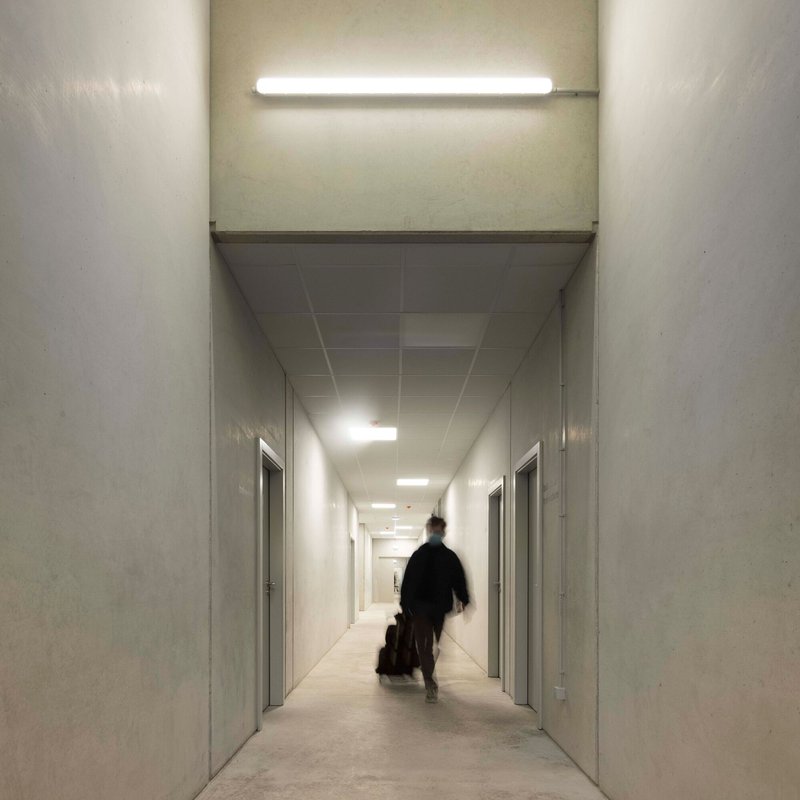 White corridor with fluorescent lighting and figure with luggage walking toward tiled ceiling terminus