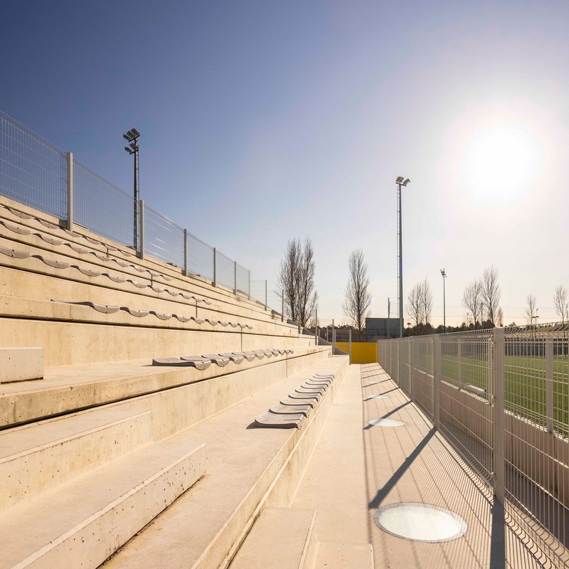 Concrete stepped seating with drainage channels beside a wire mesh fence under clear afternoon light
