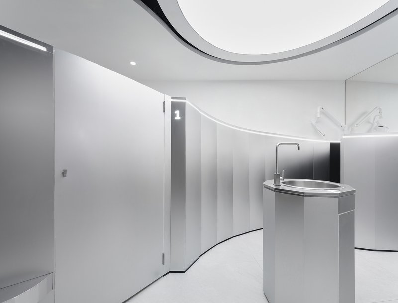 Cylindrical handwashing station with curved white cabinetry and toilet stall partitions under recessed lighting