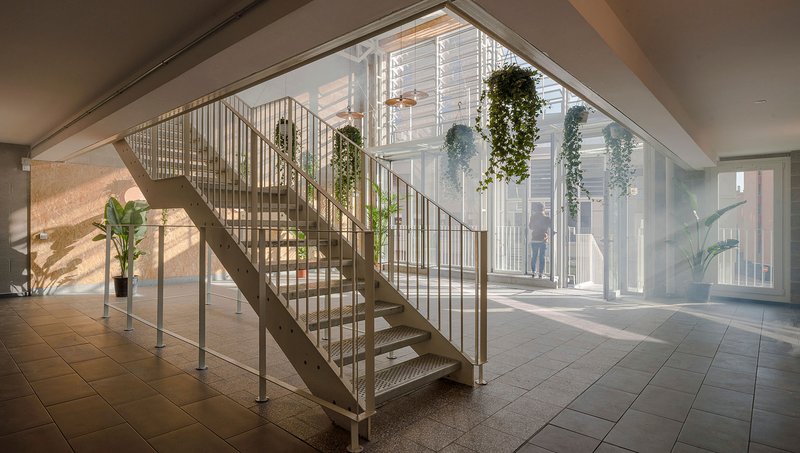 Metal staircase with vertical rod railings rising through a skylit lobby with potted plants and sunlight