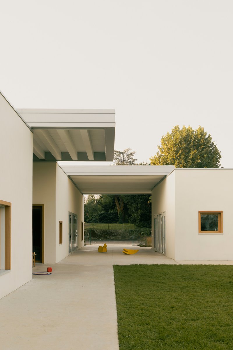 Covered passageway with exposed beam ceiling connecting white volumes across a courtyard in autumn