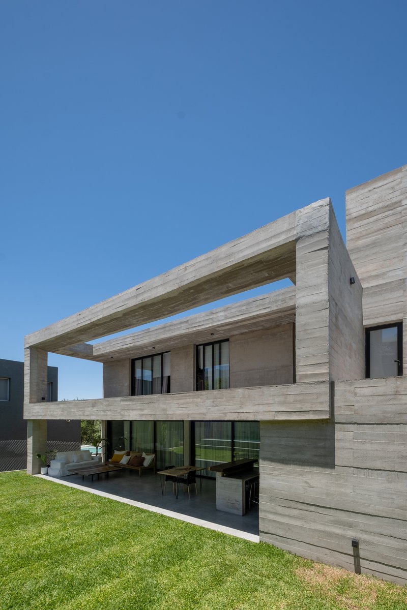 Board-formed concrete facade with cantilevered beams framing a recessed terrace above a sloped lawn