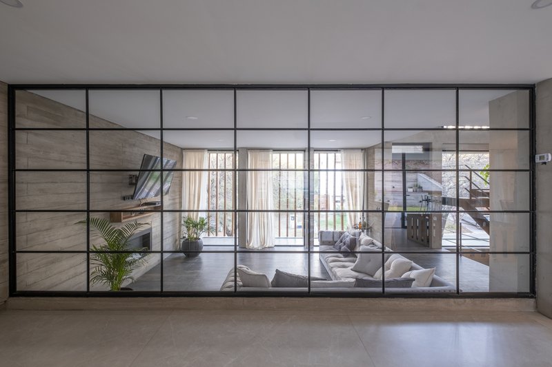 Steel-framed glass partition wall separating a hallway from a sunlit living room with curtains