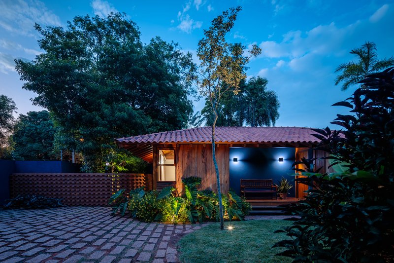 Timber-clad entrance facade with terracotta tile roof illuminated at dusk among garden trees