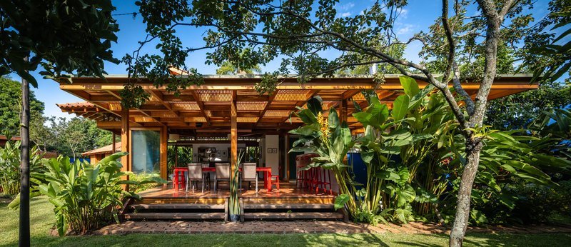 Front elevation of open pavilion with exposed timber rafters and red furniture under tree canopy