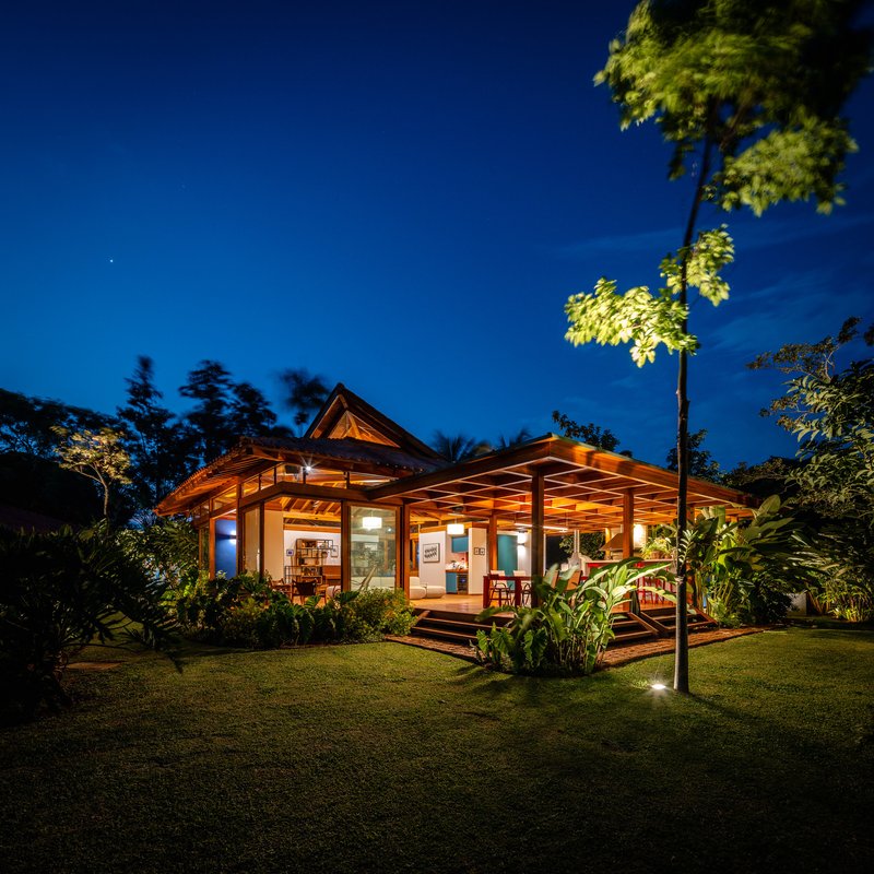 Single-story pavilion with pyramidal roof and timber pergola viewed across lawn under evening sky