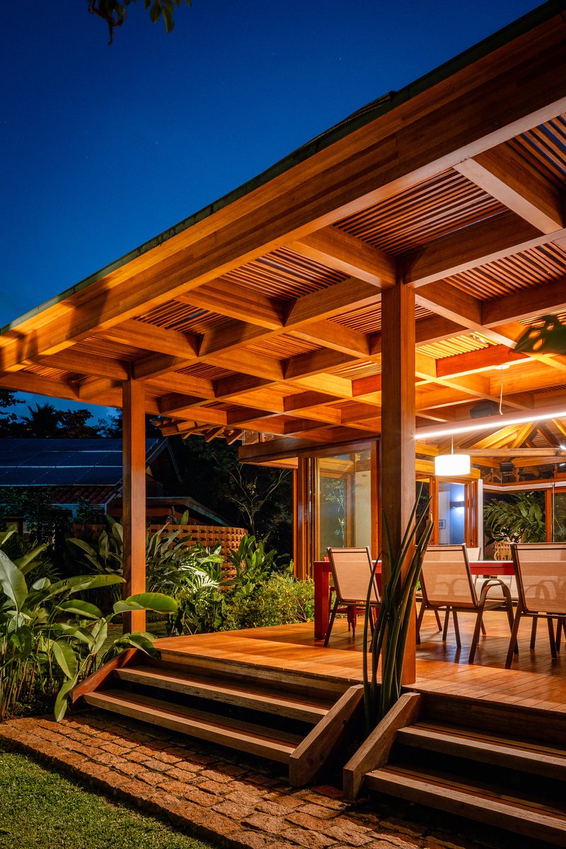 Detail of layered timber roof structure and stepped deck with chairs under illuminated eave