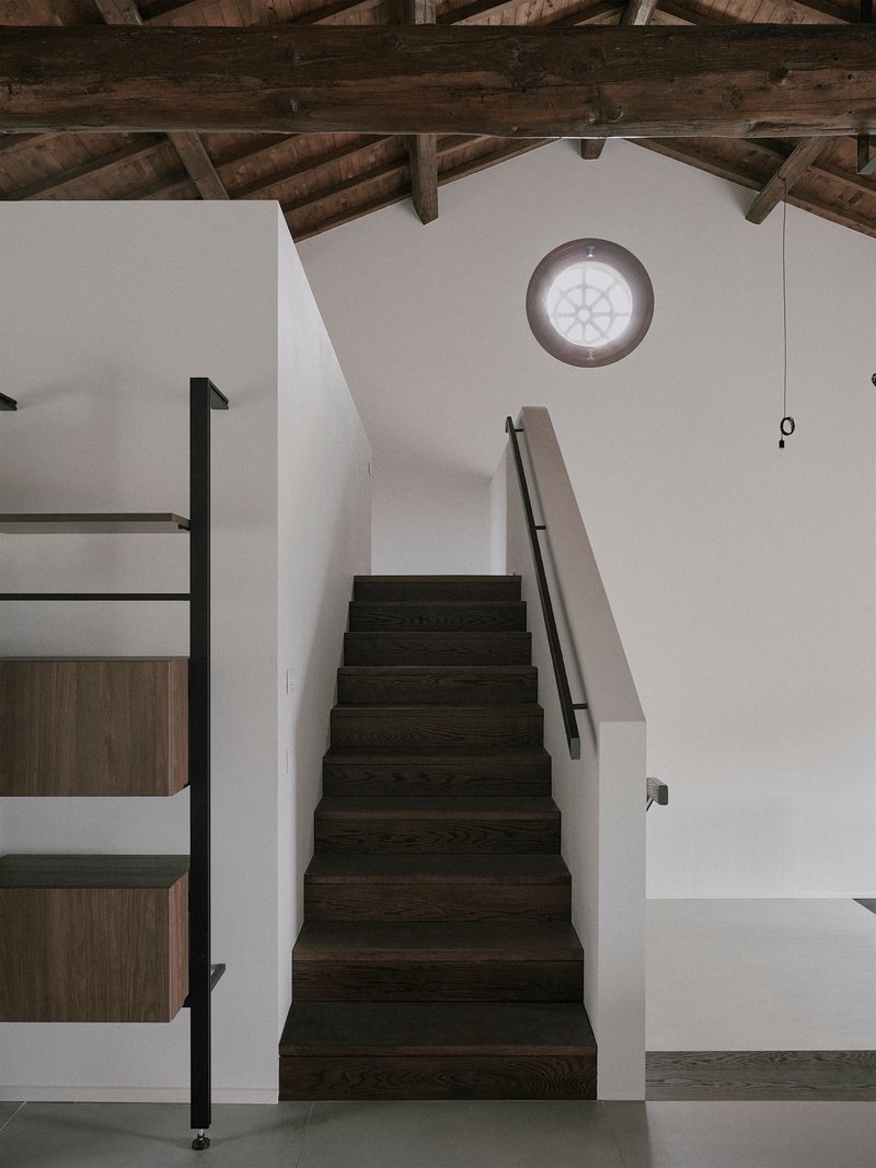 Dark timber staircase with metal handrail rising beneath exposed roof beams and a circular window