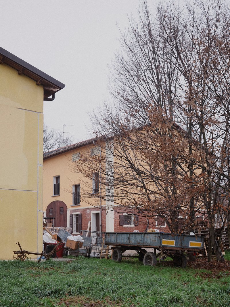 Yellow-rendered building volumes with bare deciduous trees and farm trailer in overcast weather