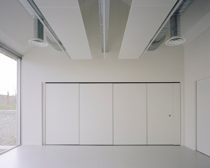 Open room with exposed ceiling ducts and perforated acoustic panels above flush storage cabinets