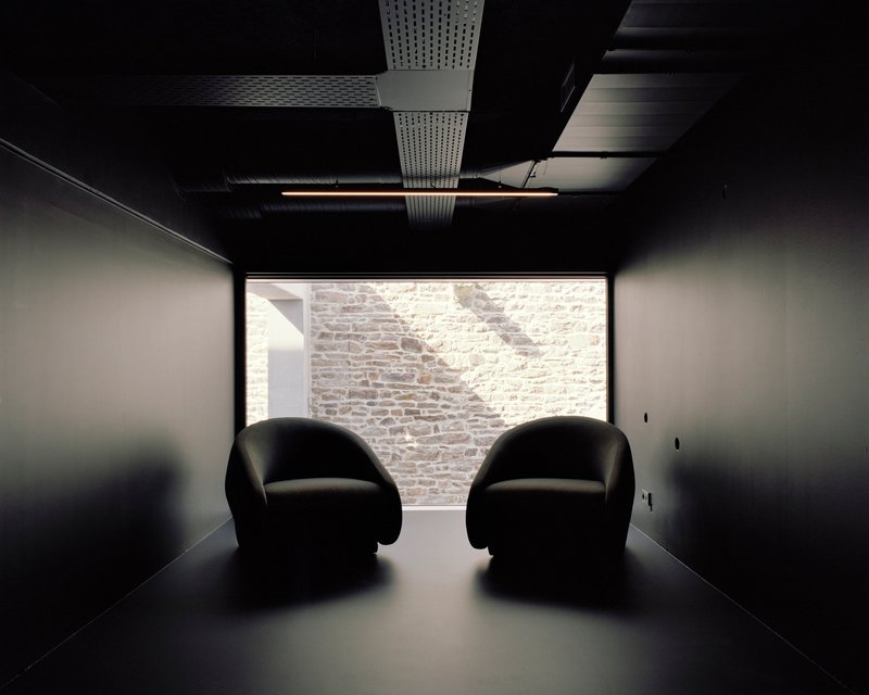 Dark room with two rounded chairs facing a window framing a sunlit stone wall