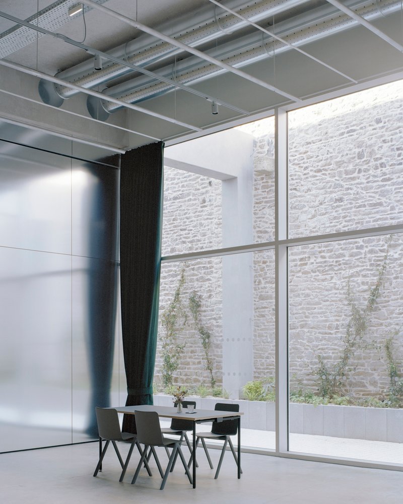 Double-height glazed wall revealing a courtyard with planted saplings against a stone masonry facade