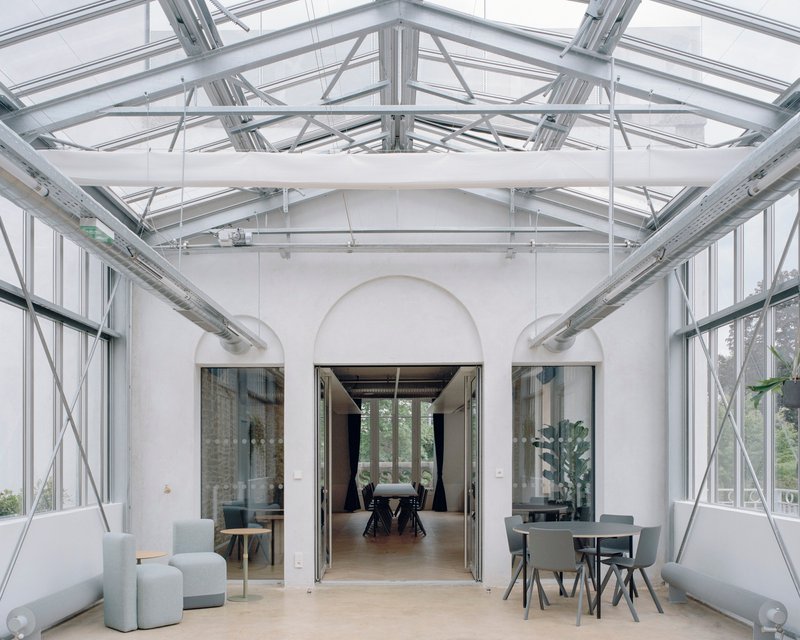 Glazed conservatory space with steel truss roof, arched opening and scattered seating under natural light