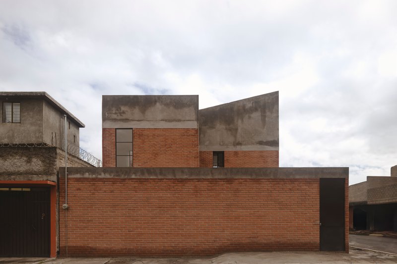 Street facade with stepped concrete volumes rising above a continuous red brick wall under cloudy sky