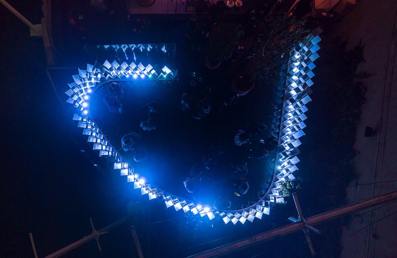 Overhead view of the horseshoe-shaped glowing pavilion with visitors moving through the courtyard