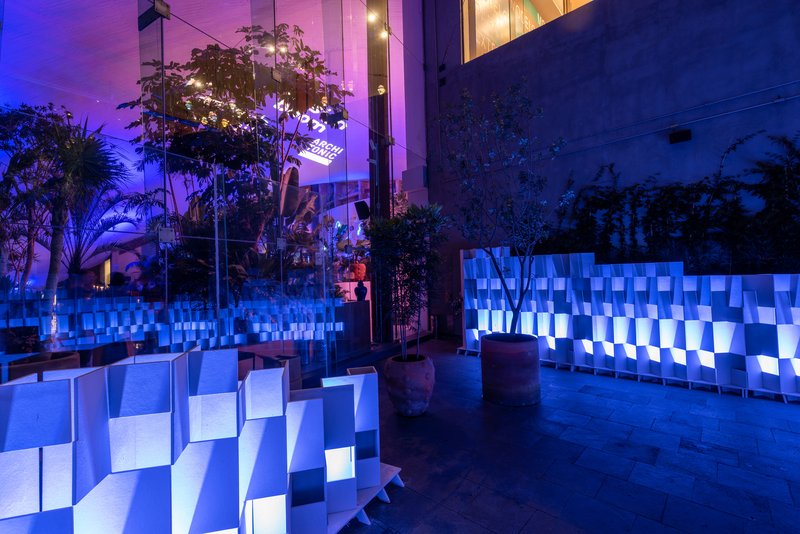 Courtyard with backlit folded seating and partition walls surrounded by potted trees under violet evening light
