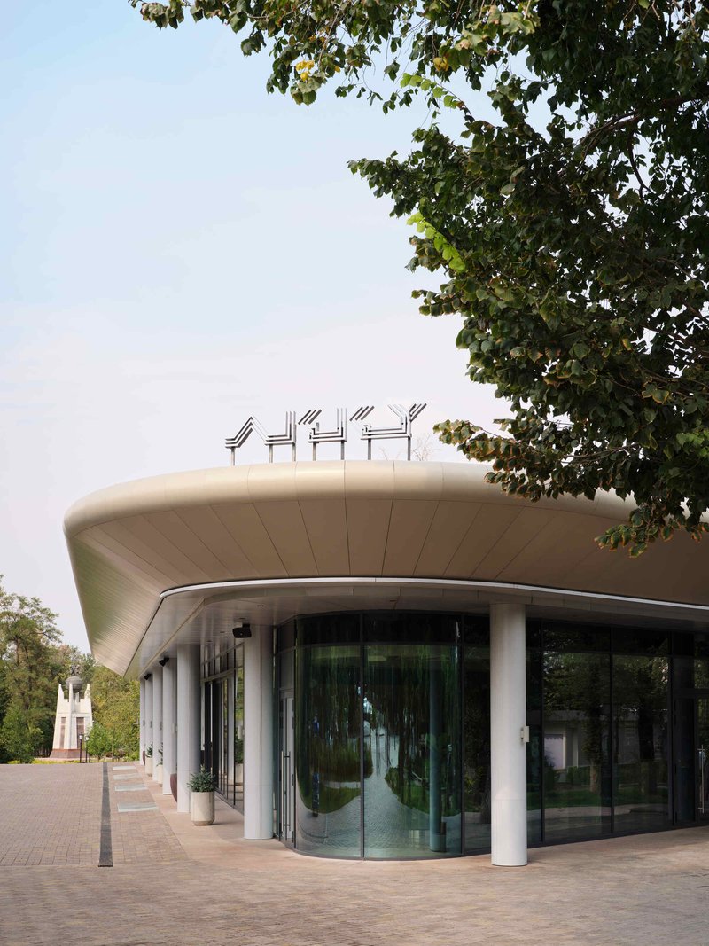 Curved facade with white columns and glazing beneath an overhanging roof framed by tree canopy