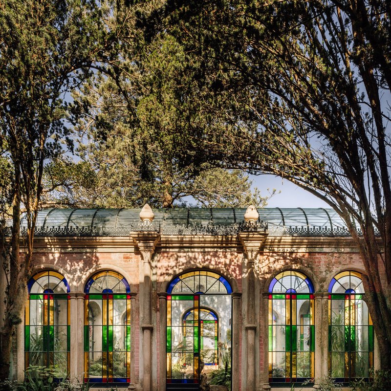 Row of arched windows with colorful stained glass beneath a barrel vault roof under mature tree canopy
