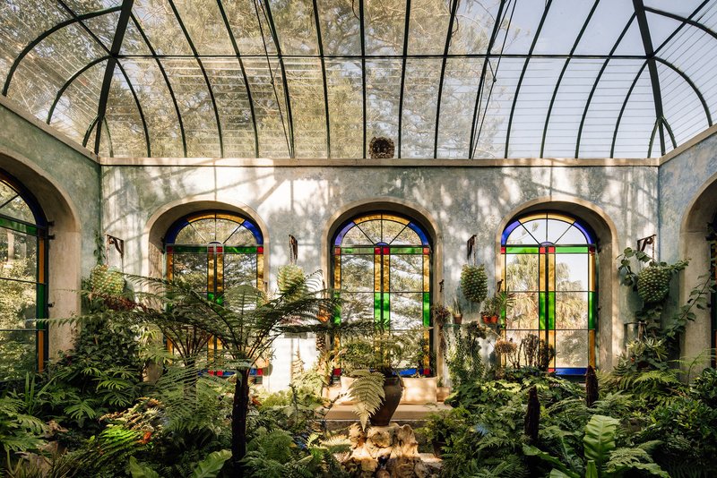 Interior conservatory with arched glass roof and stained glass windows framing potted plants and stone fountain