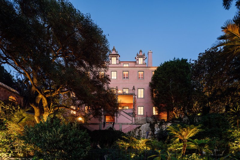 Pink rendered facade with symmetrical chimneys framed by palms and trees at dusk