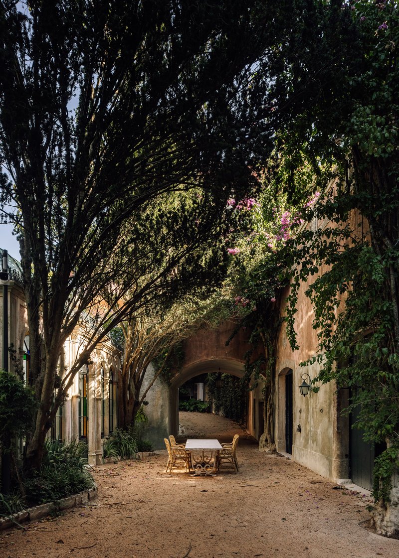 Gravel courtyard with wicker furniture under vine-covered arches and overhanging tree canopy