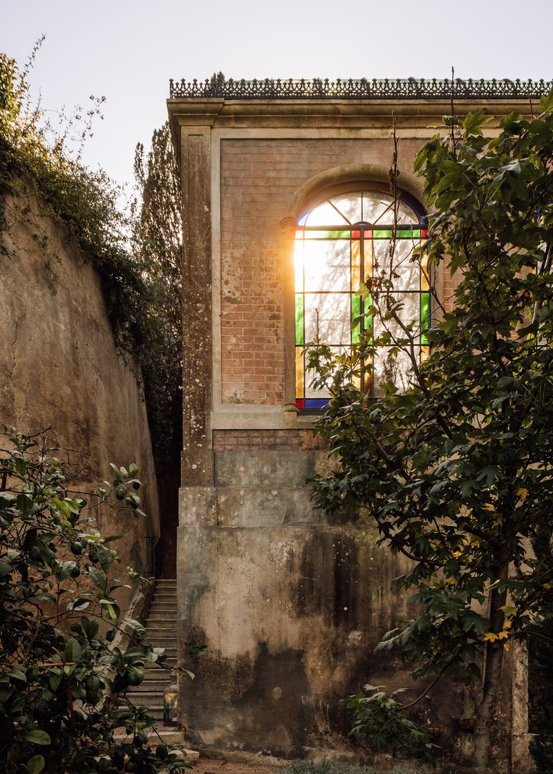 Weathered brick pavilion with arched stained glass window illuminated at dusk amid climbing vegetation