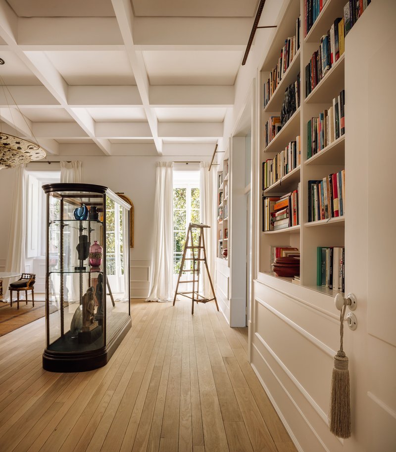 Corridor with coffered ceiling, built-in bookshelves, glass vitrine, and wooden ladder beside tall windows