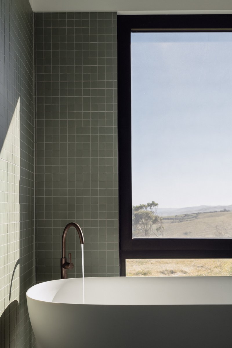 Freestanding white bathtub beneath black-framed window with green square tile wall in morning light