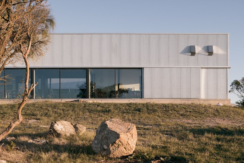 Corrugated metal facade with floor-to-ceiling glazing on a hillside at golden hour